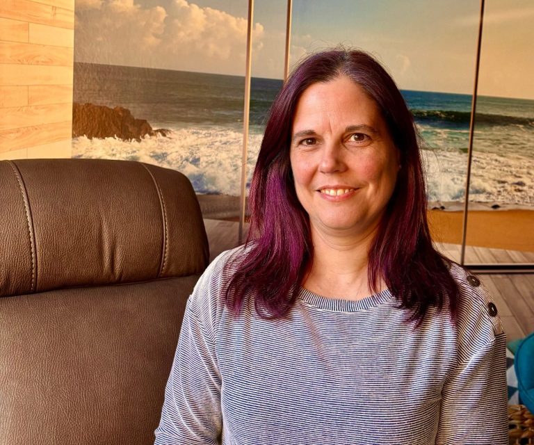 Frau mit violetten Haaren und Brille, lächelt vor einem braunen Sessel und einem verschwommenen Hintergrund.